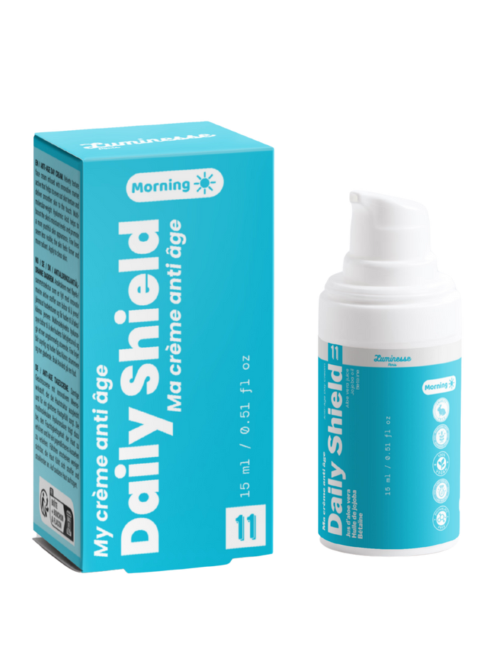 Packshot de Daily Shield n°11 de Luminesse Paris avec son étui turquoise et son flacon pompe blanc de 15 ml. La boîte et le flacon sont présentés côte à côte sur fond transparent ; le packaging indique une crème anti-âge pour le matin et mentionne notamment jus d’aloe vera, huile de jojoba et bétaïne.